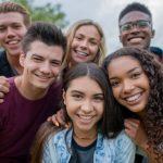 A large group of teens huddle in closely together as they casually pose for a portrait.  They are each dressed casually and are smiling as they enjoy time together outside.